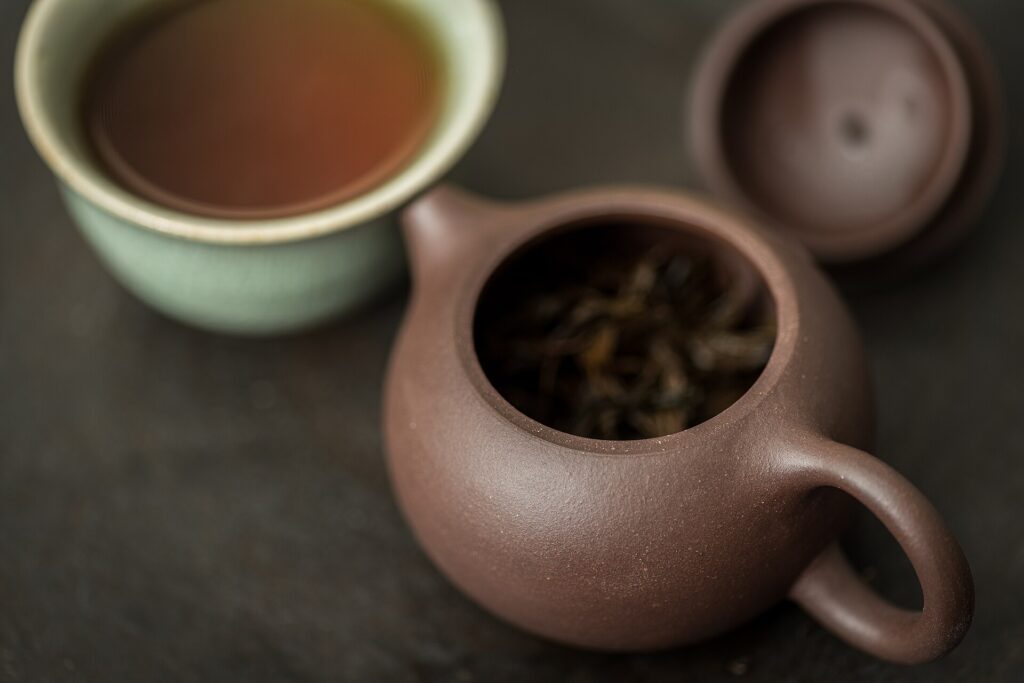 A small brown Yixing clay teapot holding dark lapsang souchong leaves, with a cup of tea behind.