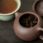A small brown Yixing clay teapot holding dark lapsang souchong leaves, with a cup of tea behind.