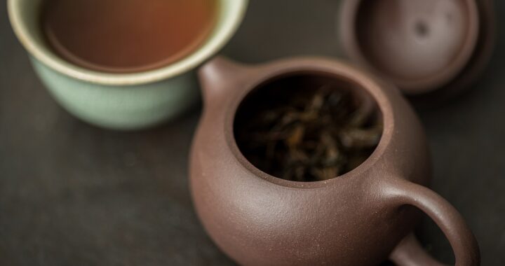A small brown Yixing clay teapot holding dark lapsang souchong leaves, with a cup of tea behind.
