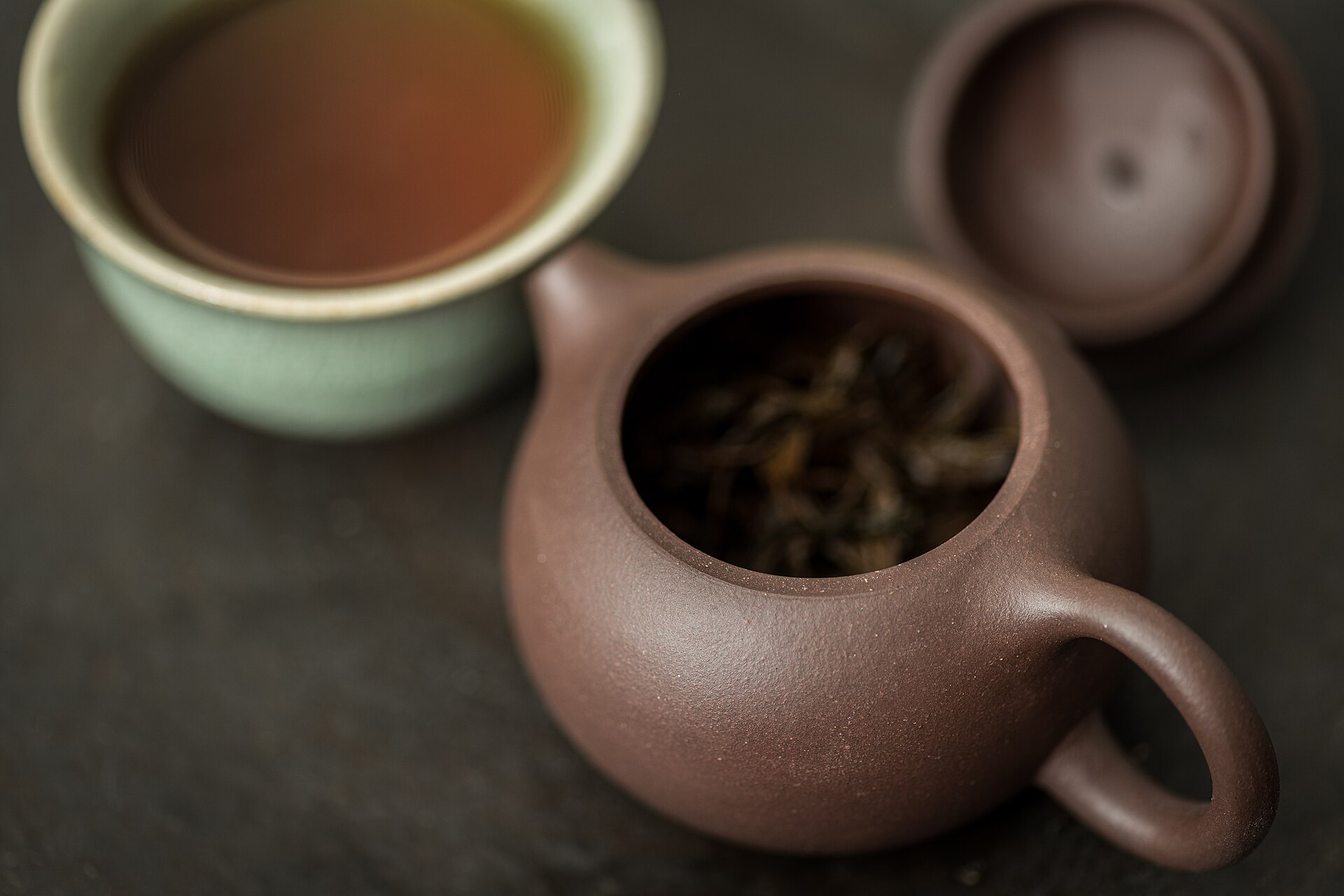 A small brown Yixing clay teapot holding dark lapsang souchong leaves, with a cup of reddish tea behind it.
