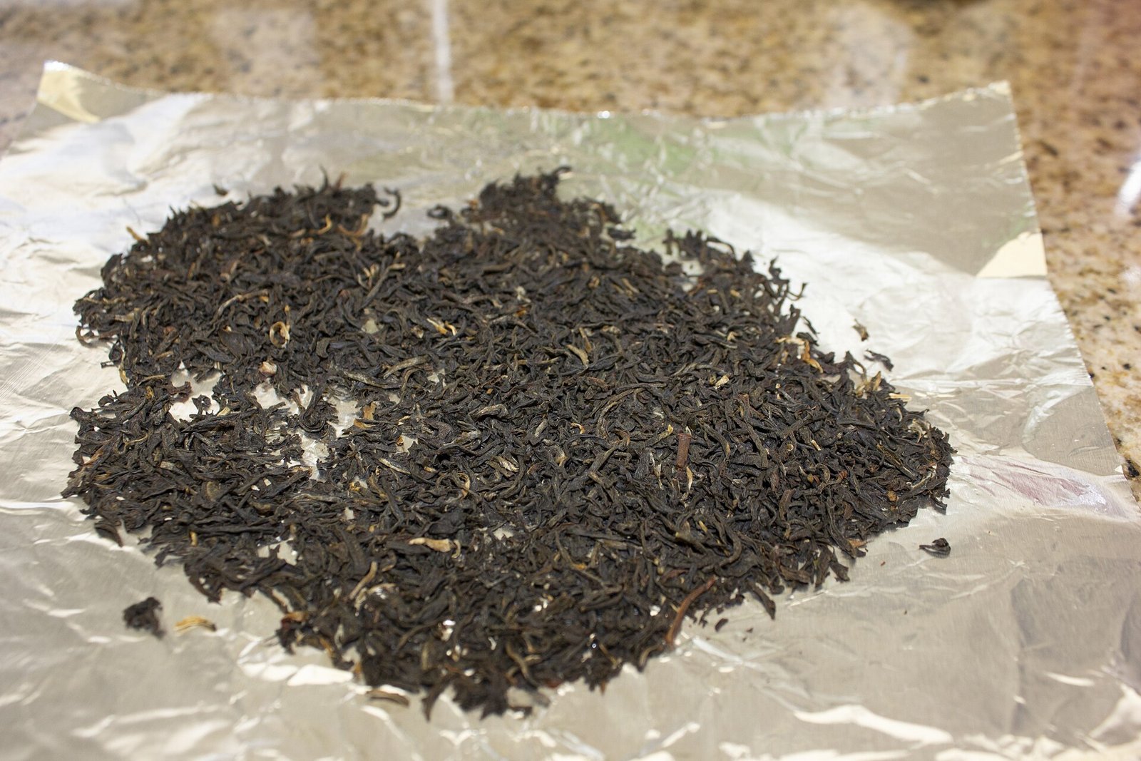A pile of dark, twisted loose-leaf Assam black tea with a few golden tips, spread on aluminium foil.
