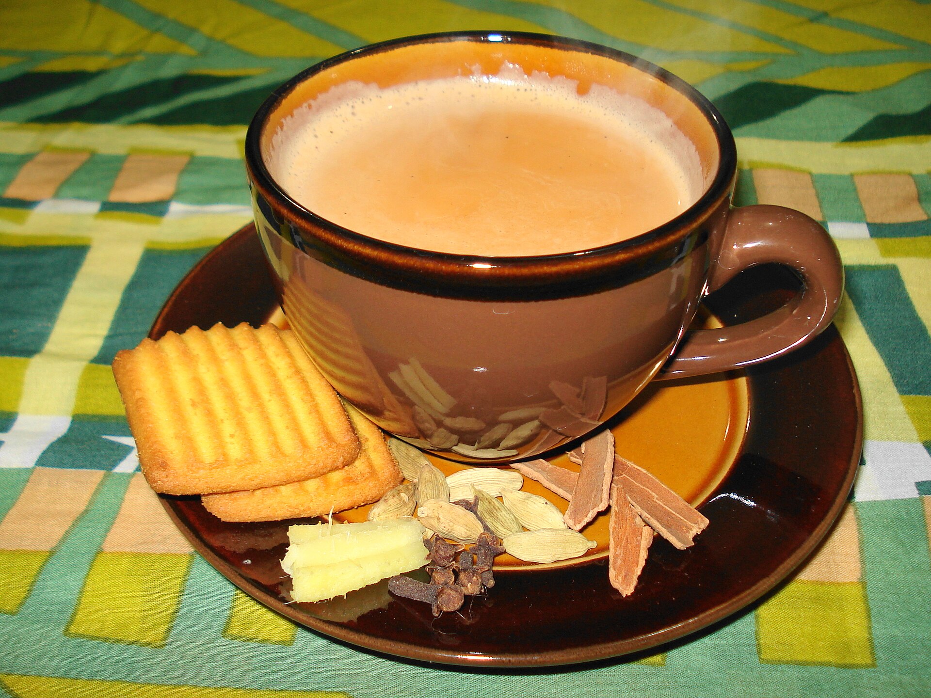 A brown cup of milky masala chai on a saucer with crackers, cinnamon sticks, cardamom pods and ginger on a patterned tablecloth.