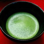 A black bowl of bright green, frothy whisked matcha on a red tray.