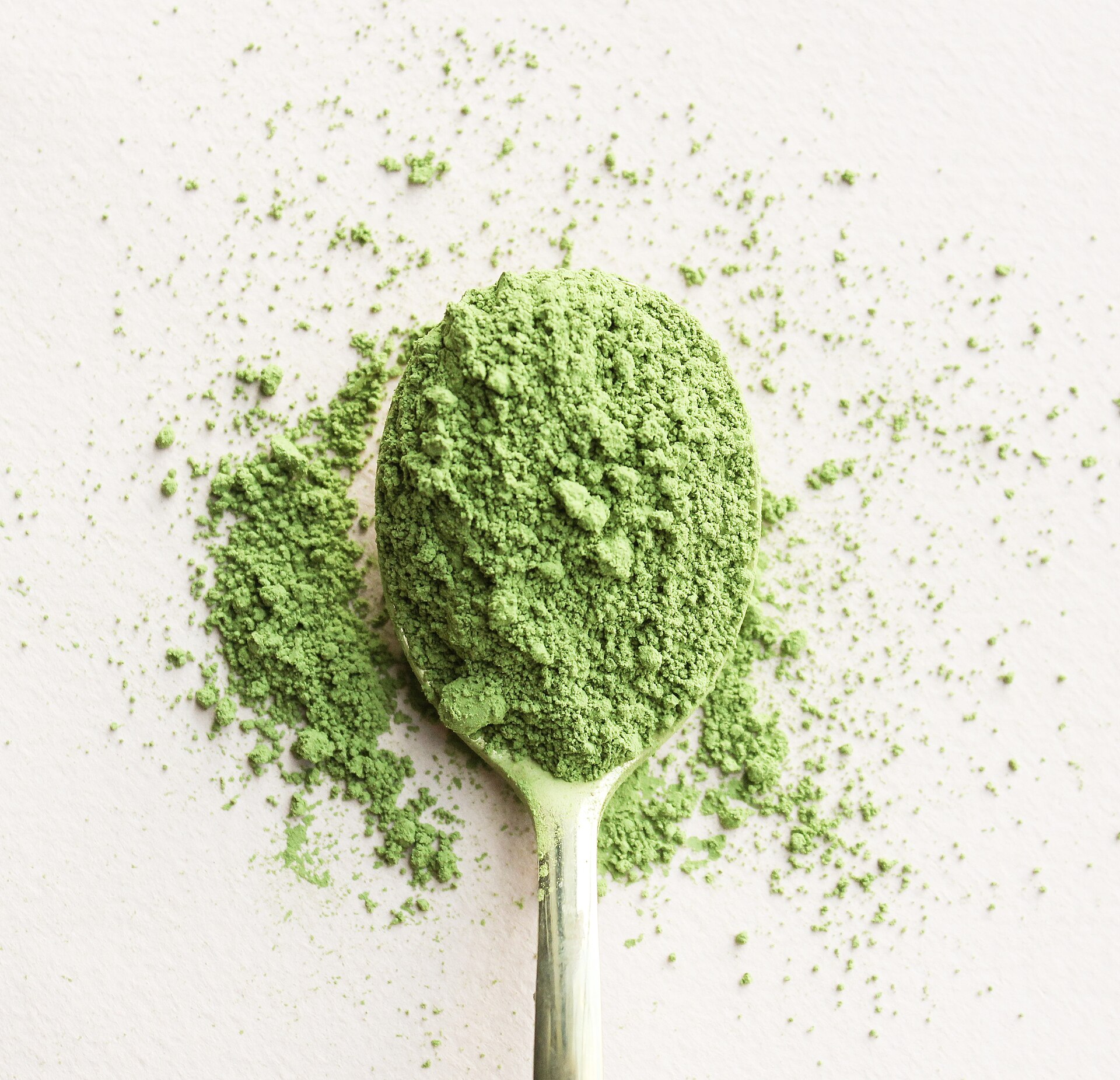 A heaped silver spoon of bright green matcha powder on a white surface, with loose powder scattered around it.