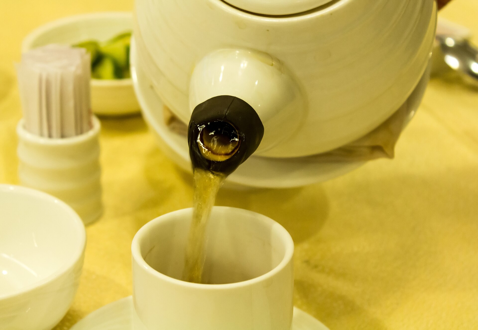 Pale golden oolong tea being poured from a cream teapot into a small white cup on a yellow tablecloth.