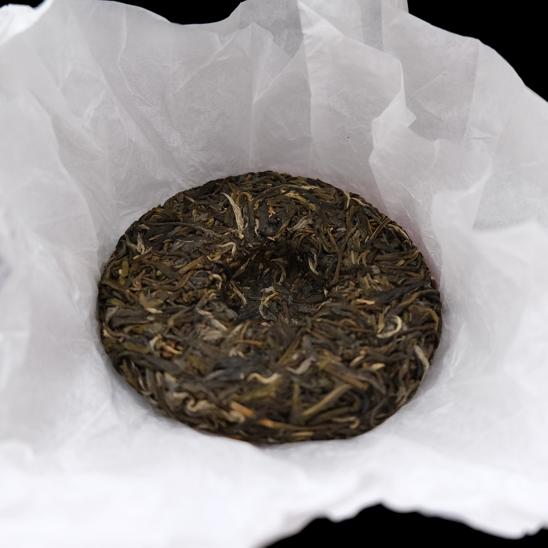 A round, flat cake of raw pu-erh tea made of pressed twisted green and silver leaves, sitting on its opened white paper wrapper.