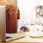 A white cup of tea with a tea bag tag over the rim, on a wooden desk beside an open laptop and a leather bag.