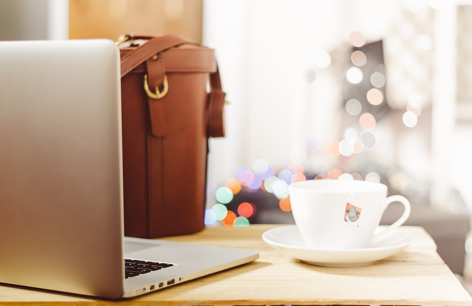 A white cup of tea with the tag of a tea bag hanging over the rim, on a wooden desk beside an open laptop and a leather bag.