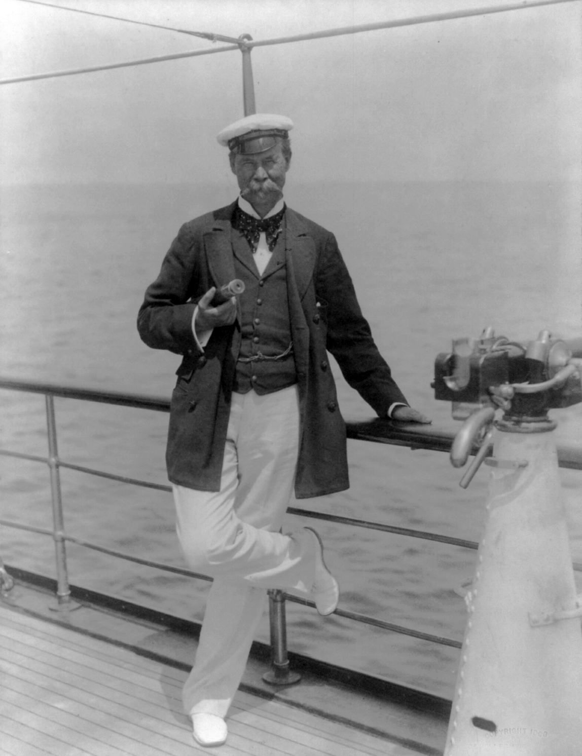 Black and white photograph of Sir Thomas Lipton standing at the rail of his yacht in a cap and light trousers.
