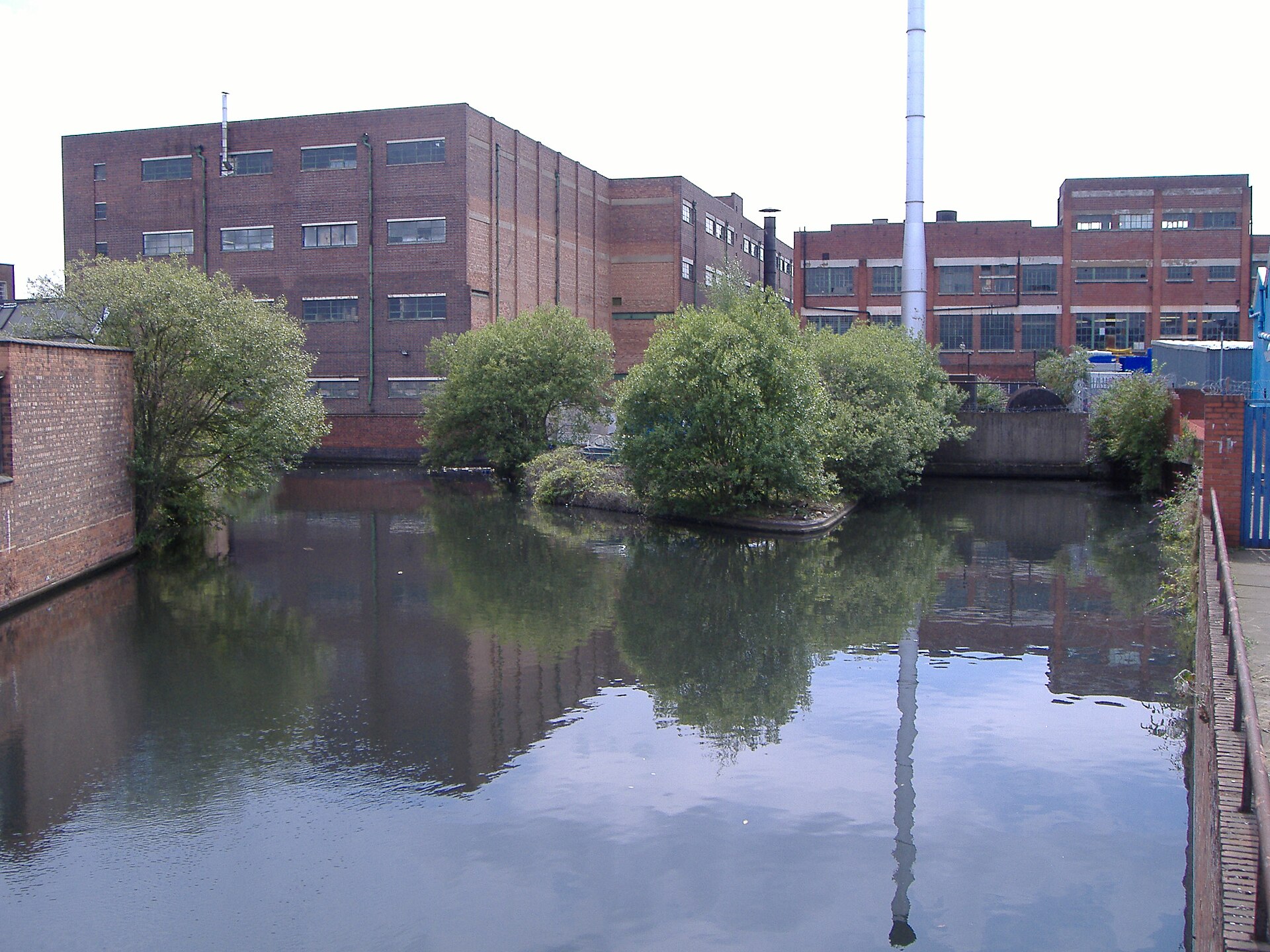 Large red-brick factory buildings with rows of windows beside a still canal basin and trees.