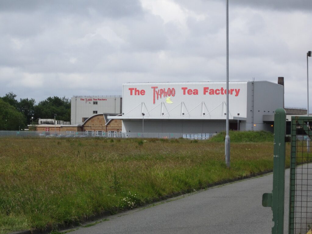 A large grey industrial building with red lettering reading The Typhoo Tea Factory, behind a field of long grass.