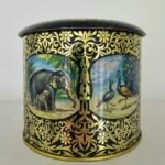A round tin tea caddy from the 1970s, black with gold floral patterns and painted panels of elephants and peacocks.