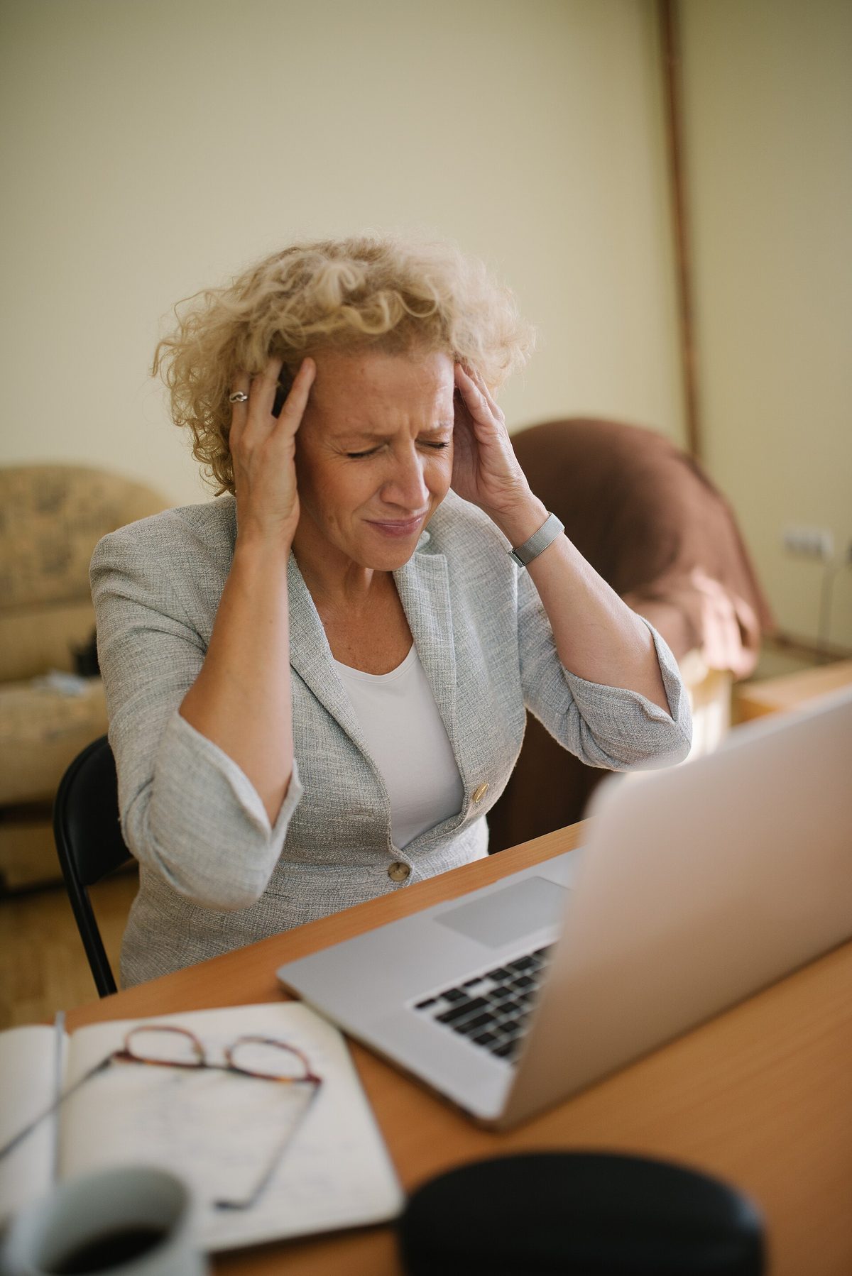 A woman sitting at a desk with a laptop, eyes closed, pressing her fingers to her temples.