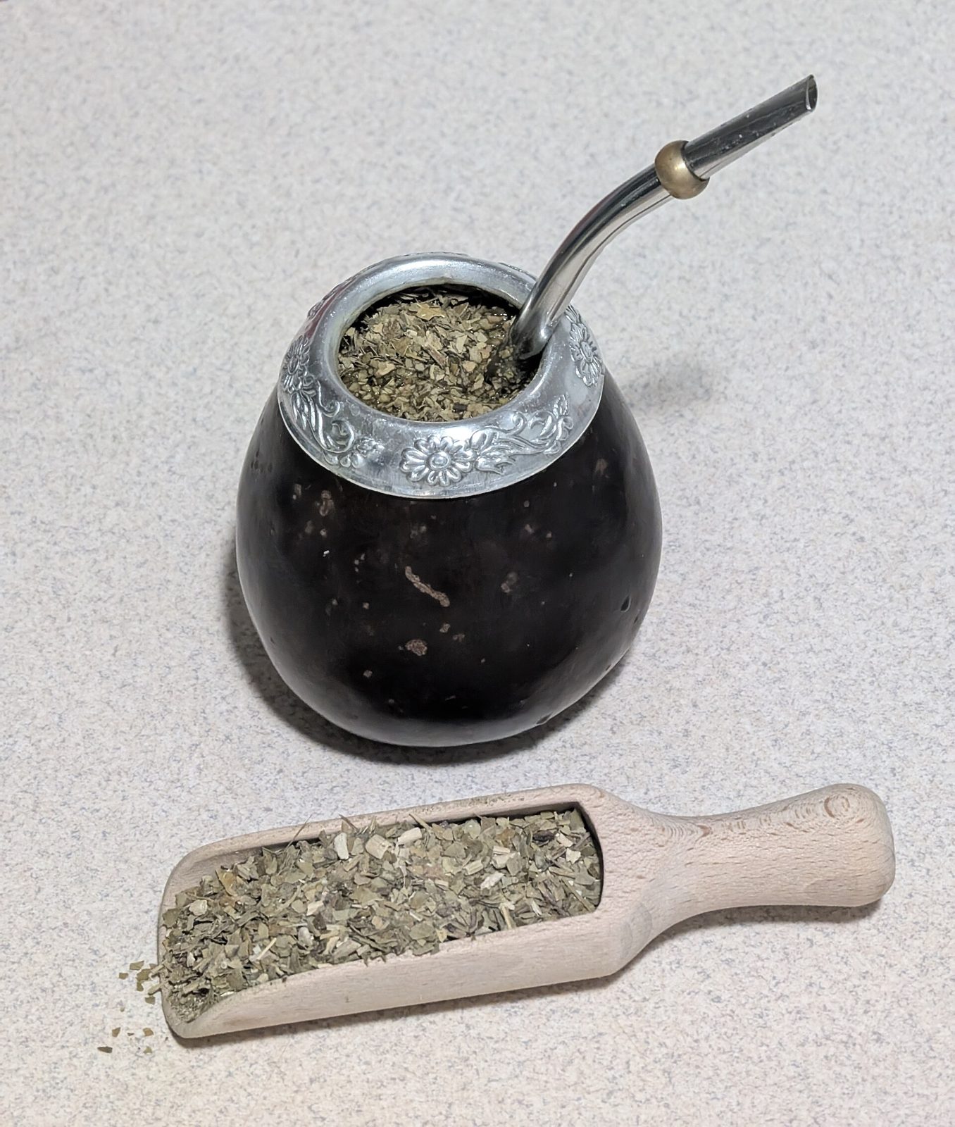 A dark calabash gourd with an engraved metal rim, filled with dry yerba mate and a metal straw, next to a wooden scoop of loose yerba mate.