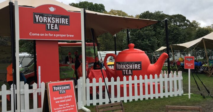 A red Yorkshire Tea stand with a giant red teapot, white picket fence and signs reading Come on in for a proper brew, on grass in Harrogate.